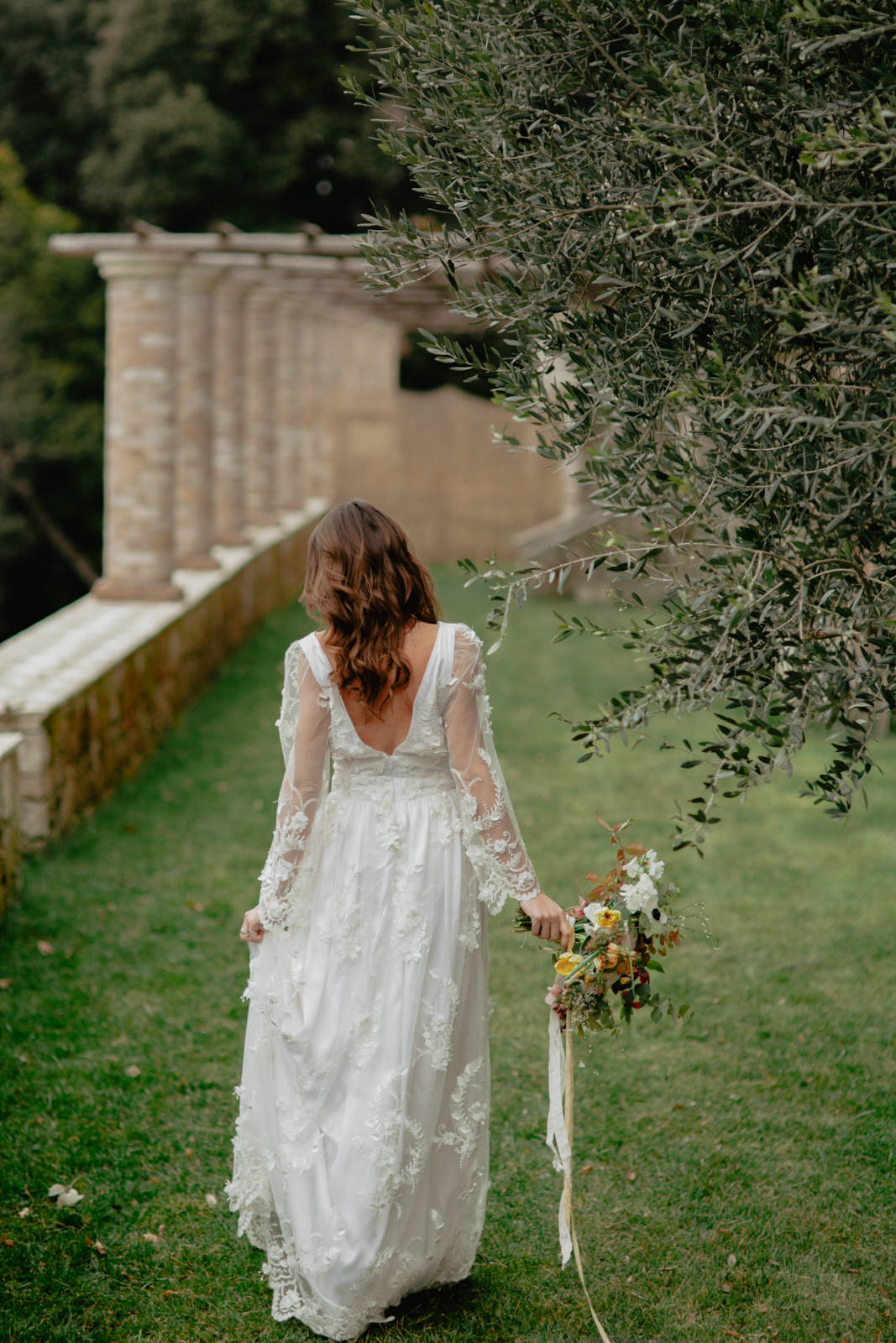 A woman in a long, white lace wedding dress carries a floral bouquet, walking gracefully on green grass. Near a stone structure with arches and a drooping tree, the scene is serene and picturesque—perfect for an intimate wedding photographer in Tuscany.