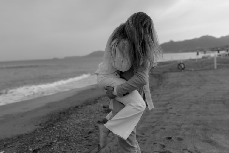 A black and white image captures a couple embracing playfully on a beach in Italy, reminiscent of a serene wedding at Borgio Verrezzi. The woman is being lifted and carried by the man as ocean waves gently crash on the shore, with mountains visible in the distance.