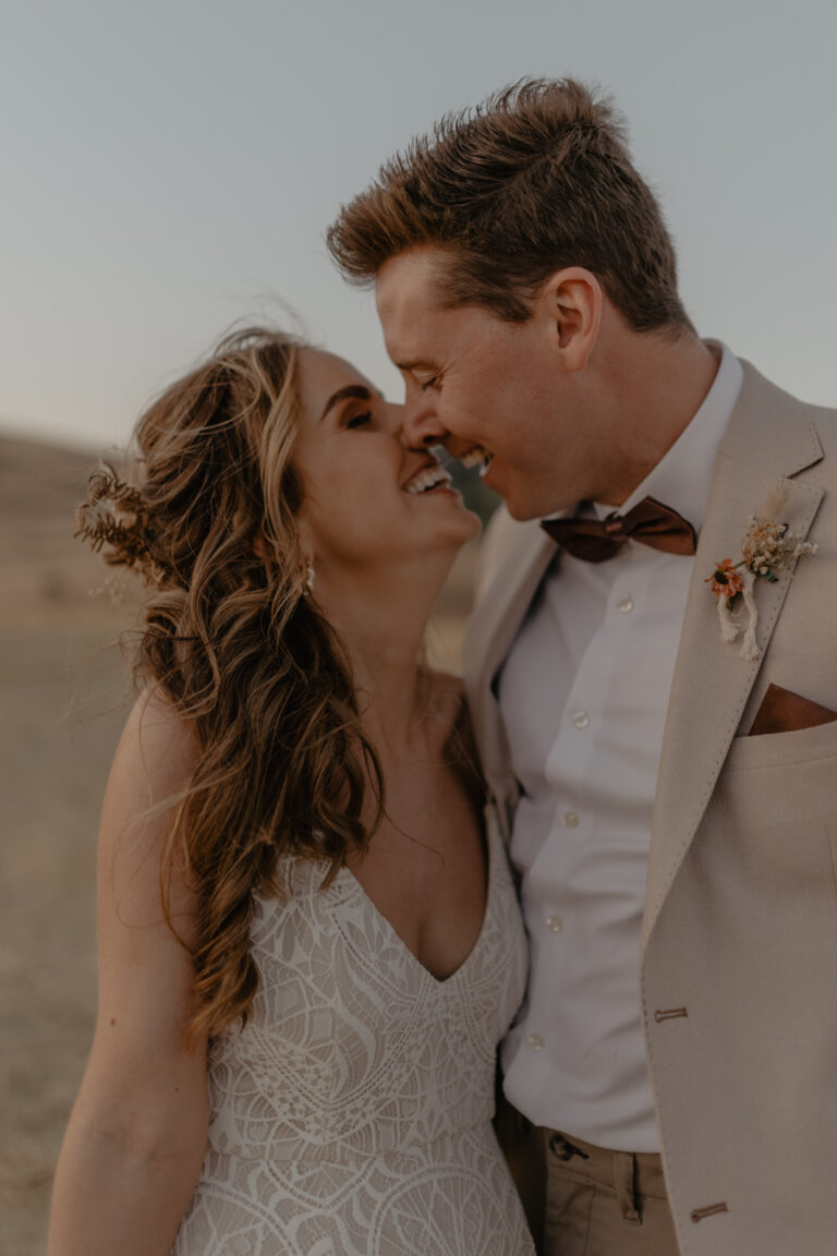 At a Boho wedding at Masseria Susafa, captured by Federica Cavicchi, a couple stands in wedding attire amidst an enchanting outdoor setting. Smiling and touching noses affectionately, the bride dazzles in lace while the groom looks dapper in his beige suit against the blurred natural backdrop.