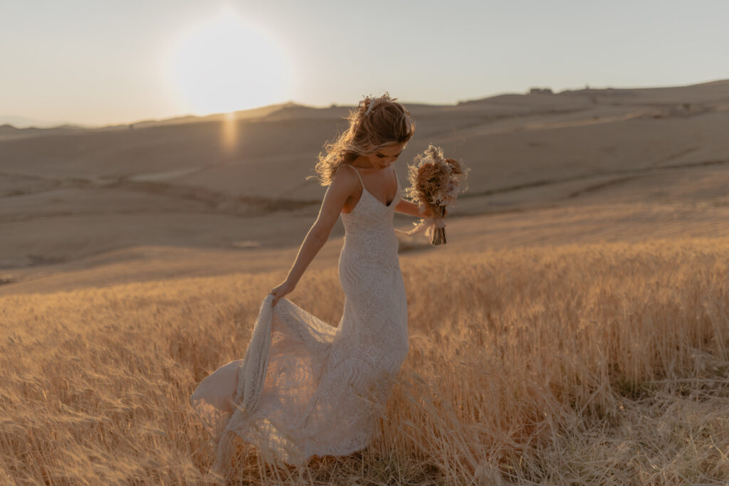 A woman in a white lace dress holds a bouquet as she strolls through a golden wheat field at sunset, capturing the boho wedding essence at Masseria Susafa. The sun casts a warm glow over the landscape, enhanced by Federica Cavicchis touch, creating a serene and romantic atmosphere.
