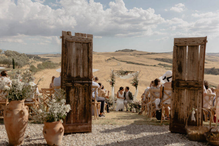 A couple is getting married outdoors in a picturesque setting with rolling hills, celebrating a sustainable and intimate wedding at The Lazy Olive. Guests dressed in light colors sit on wooden chairs, surrounded by rustic wooden doors and decorative vases filled with vibrant flowers.
