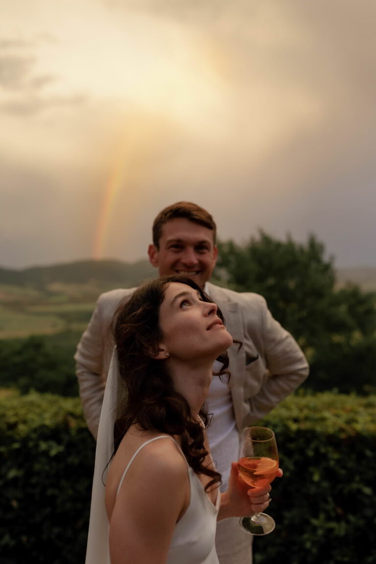 Amidst the lush greenery of Borgo di Castelvecchio, Tuscany, a bride in a white dress gazes up at the sky, wine glass in hand. Her groom, clad in a beige suit, stands behind her with a smile. A rainbow graces the cloudy sky—captured beautifully by Federica Cavicchi Photography.
