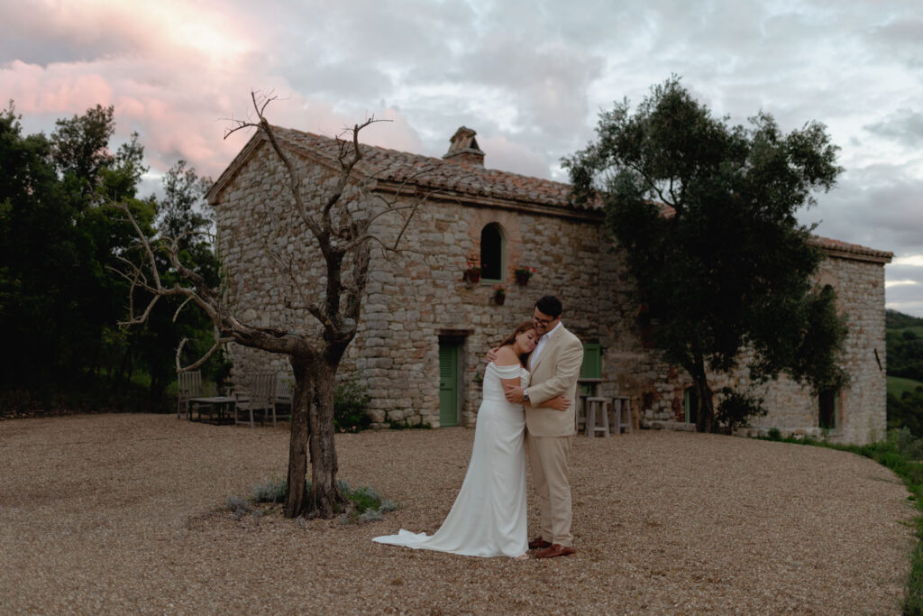A couple embraces in front of a rustic stone house at Tenuta di Murlo, surrounded by trees and a cloudy sky at sunset. She wears a white dress, he wears a beige suit, standing on gravel next to a leafless tree. Captured by Federica Cavicchi, the atmosphere is serene and romantic.