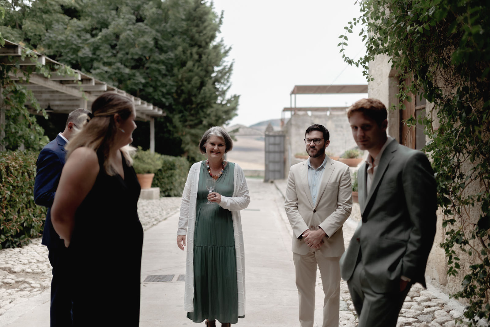 A Romantic Destination Wedding at Masseria Susafa 57 A group of people stands outside on a stone path surrounded by greenery. One woman in a green dress and white cardigan is facing the camera, while a man in a beige suit looks on. Others are chatting in the background. The setting appears casual and relaxed.