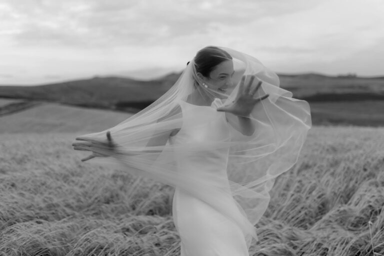 A bride in a white dress and veil joyfully dances in a field of tall grass. The veil flows elegantly as she moves, and the background shows rolling hills under a cloudy sky. The scene is in black and white.