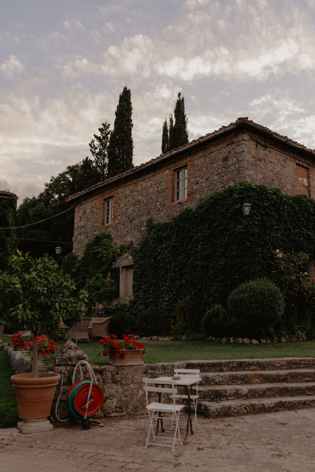 Timeless and Elegant Tuscan Wedding at Borgo Stomennano: 103 A rustic stone house covered in ivy stands under a cloudy sky. In front, a patio features a white table and two chairs, potted plants, and a coiled garden hose. Tall cypress trees rise in the background.