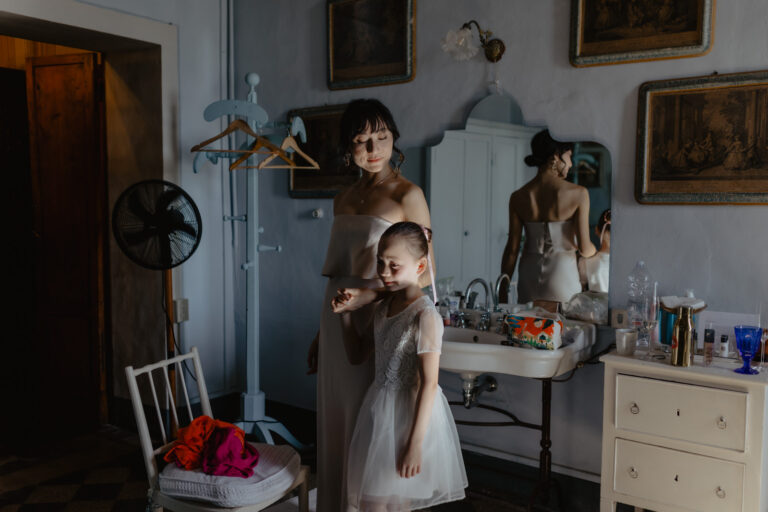 A woman in a strapless dress stands beside a young girl in a white dress inside a dimly lit room. They are near a vintage sink with a mirror and various toiletries. A chair and coat rack are visible nearby.