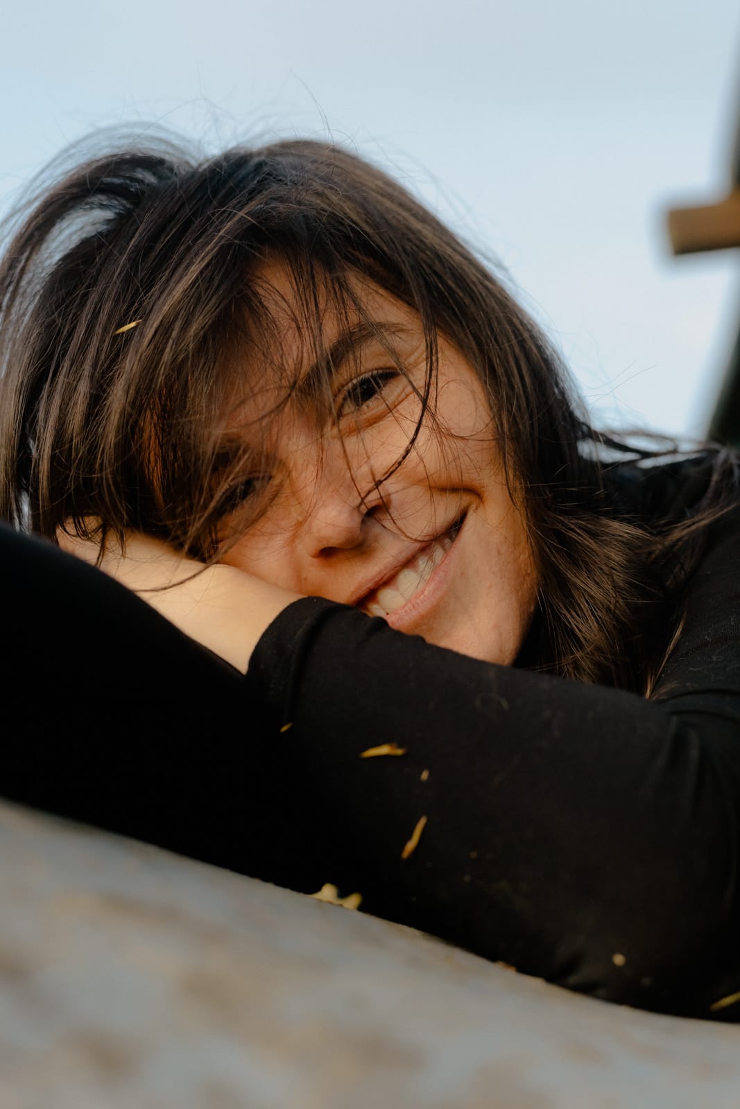 A young woman with long dark hair smiles warmly, lying on her side with her head resting on her arm. She is wearing a black top and appears relaxed and happy, with soft natural light illuminating her face.