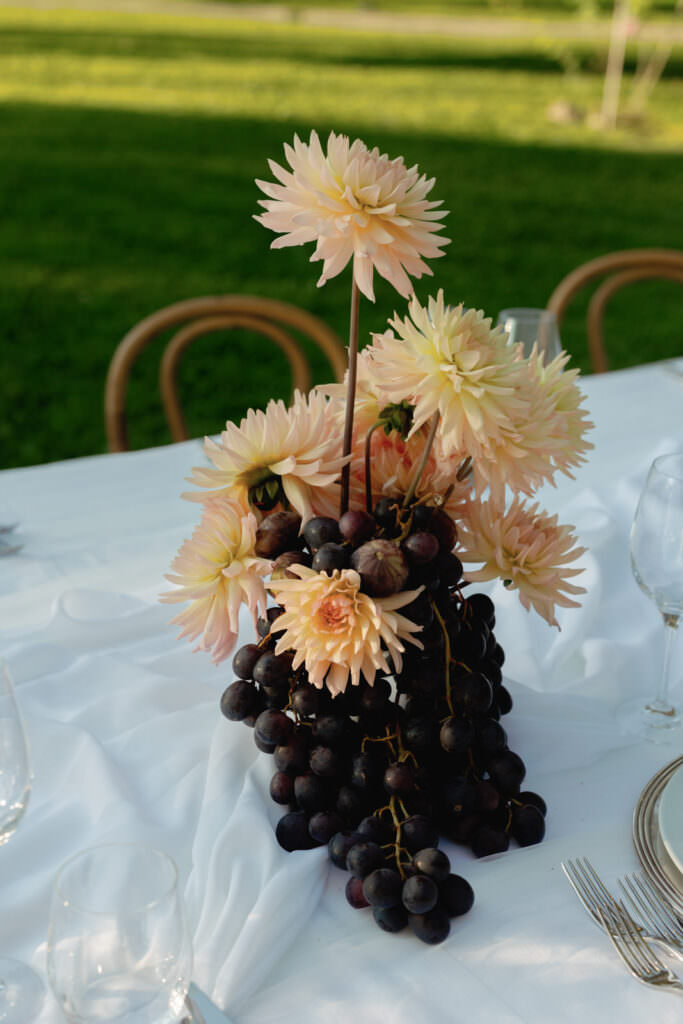 A Three-Day Destination Wedding in Tuscany 2 A centerpiece of pale pink dahlias and a cluster of dark grapes arranged on a white tablecloth, with empty wine glasses and cutlery nearby, set outdoors on a green lawn.