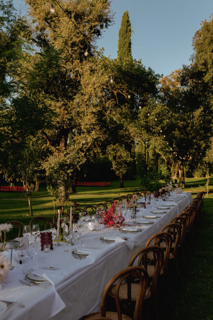 A Three-Day Destination Wedding in Tuscany 93 A long outdoor dining table with white tablecloths is set with plates, glasses, and floral centerpieces in a green, tree-filled garden at sunset. Wooden chairs line both sides of the table.