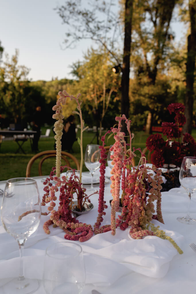 A Three-Day Destination Wedding in Tuscany 52 A white tablecloth with empty wine glasses and a centerpiece featuring cascading pink and green flowers, set outdoors in a garden with trees in the background.