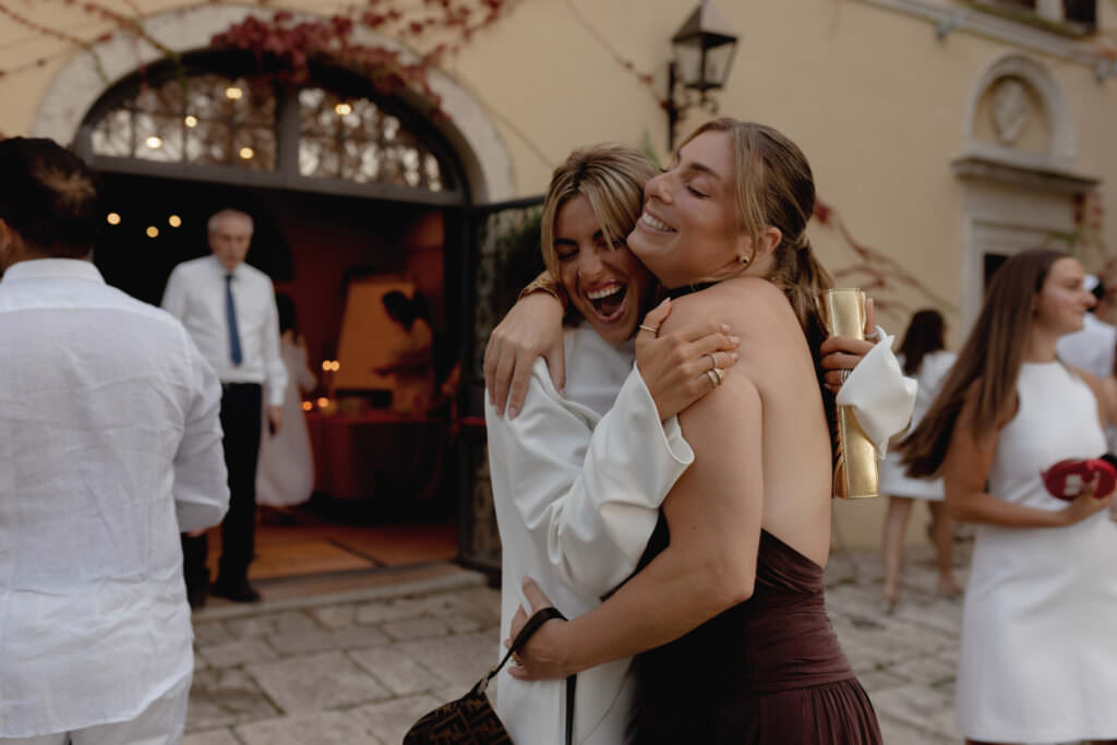 A Three-Day Destination Wedding in Tuscany 65 Two women hug and smile joyfully at an outdoor event, dressed in formal attire. Other guests in white clothing mingle in the background near a building with arched doors and ivy on the walls.
