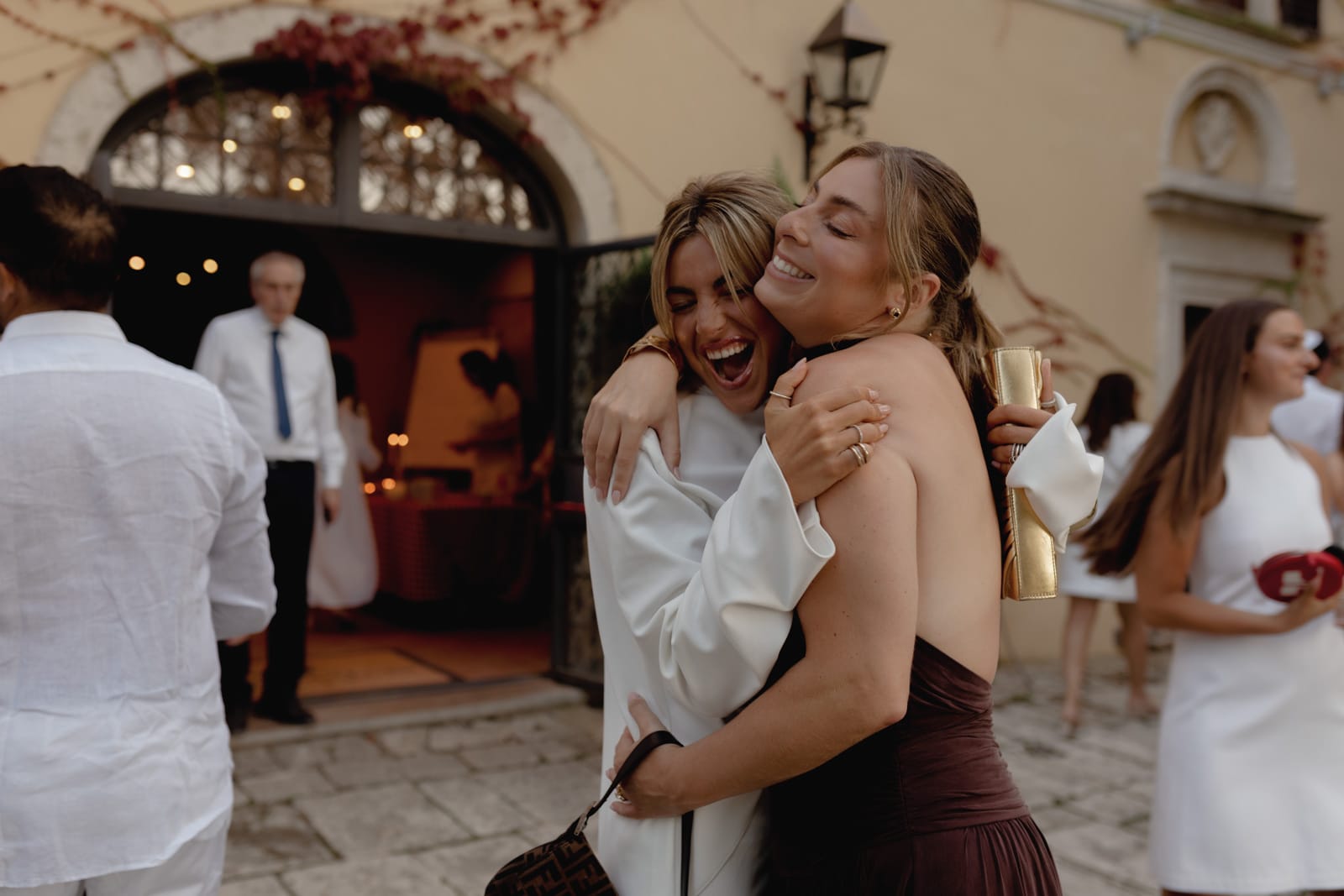 Two women hug and smile joyfully at an outdoor event, dressed in formal attire. Other guests in white clothing mingle in the background near a building with arched doors and ivy on the walls.