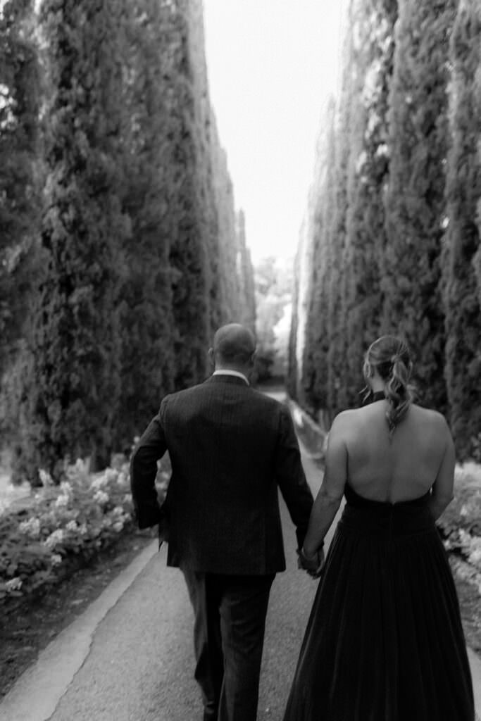 A Three-Day Destination Wedding in Tuscany 35 A couple dressed formally, holding hands, walk down a tree-lined path. The photo is black and white, with tall trees on either side, creating a romantic and serene atmosphere.