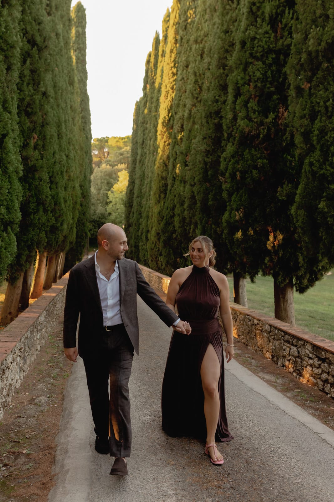 A couple walks hand in hand down a tree-lined path at sunset. The man wears a dark suit and white shirt, while the woman wears a dark dress with a high slit. Tall cypress trees and a stone wall border the path.