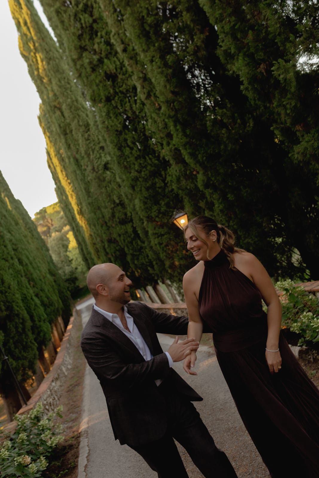 A man in a dark suit and a woman in a sleeveless dark dress walk hand in hand down a tree-lined path, smiling at each other, surrounded by tall cypress trees and greenery.