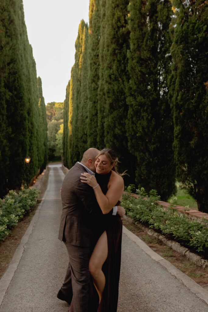 A Three-Day Destination Wedding in Tuscany 5 A couple embraces and laughs on a tree-lined path. The woman wears a black dress with a high slit, and the man wears a dark suit. Tall green trees stand on either side of the walkway in soft evening light.