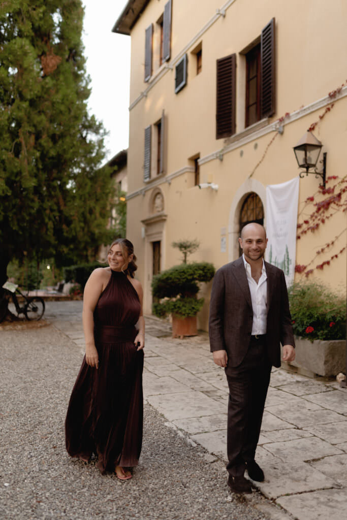 A Three-Day Destination Wedding in Tuscany 46 A woman in a dark dress and a man in a suit walk and smile outside a yellow building with open shutters and potted plants, on a stone path with greenery and a bicycle in the background.