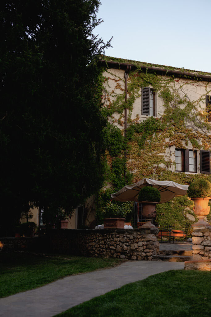 A Three-Day Destination Wedding in Tuscany 14 A stone path leads to a house covered in green ivy, with dark window shutters and a large patio umbrella shading a seating area. The scene is surrounded by lush greenery and soft evening light.