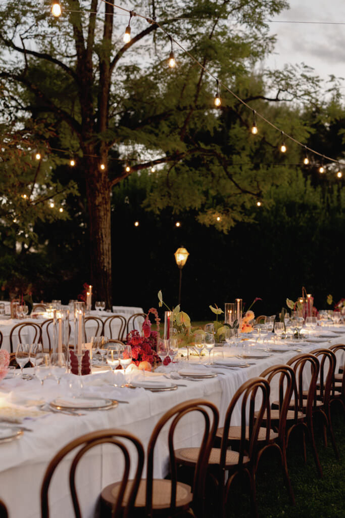 A Three-Day Destination Wedding in Tuscany 61 Long outdoor dining table set with white tablecloths, elegant place settings, candles, and floral centerpieces, surrounded by wooden chairs under string lights in a garden at dusk.