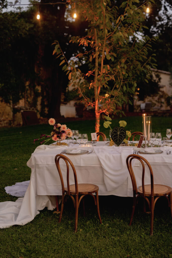 A Three-Day Destination Wedding in Tuscany 71 An elegant outdoor dinner table with a white tablecloth, wooden chairs, floral arrangements, candles, and wine glasses, set on a grassy lawn under string lights near a tree in the evening.