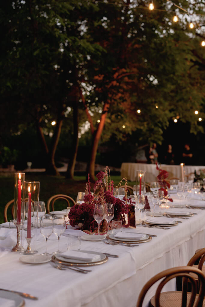A Three-Day Destination Wedding in Tuscany 12 A long outdoor dining table with a white tablecloth is set with plates, glasses, red candles, and burgundy floral centerpieces. String lights hang above, and trees and greenery surround the area at dusk.
