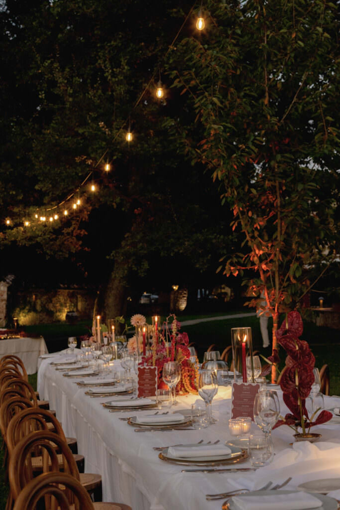 A Three-Day Destination Wedding in Tuscany 38 A long outdoor dining table decorated with white tablecloth, elegant place settings, red floral centerpieces, and lit candles, set under string lights in a garden at dusk. Wooden chairs line both sides of the table.