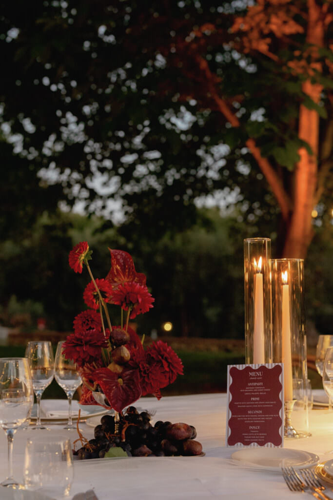 A Three-Day Destination Wedding in Tuscany 66 A white table set outdoors at dusk features a red floral centerpiece, dark grapes, a menu card, wine glasses, and two tall candles in glass holders, with trees and soft evening light in the background.