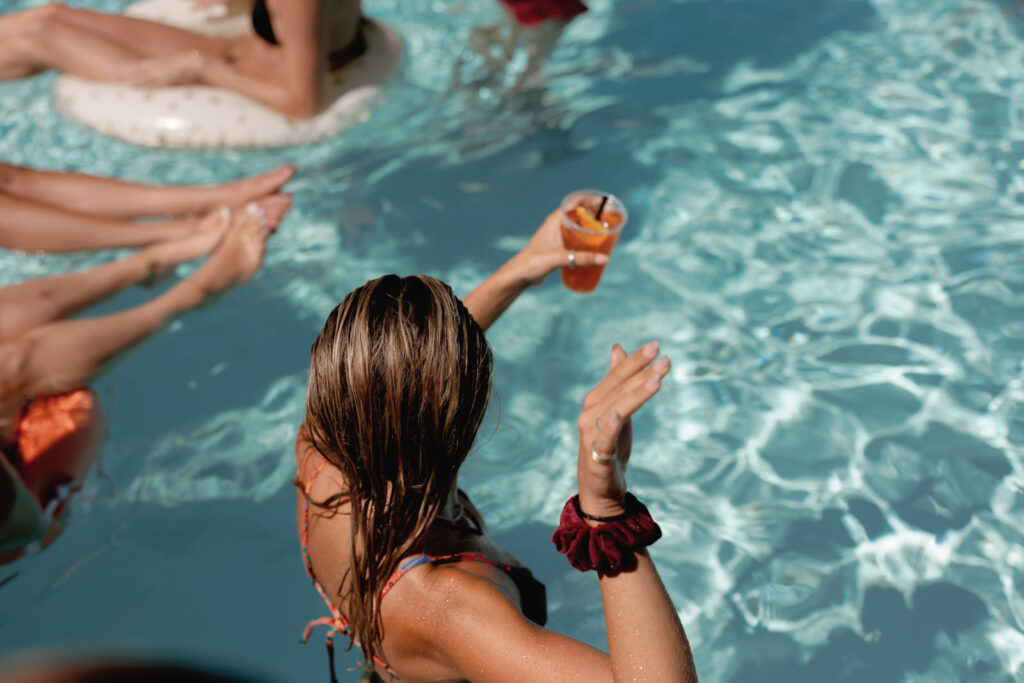 A Three-Day Destination Wedding in Tuscany 87 A person with wet hair sits at the edge of a pool holding a drink with an orange slice, surrounded by others relaxing in the water on a sunny day.