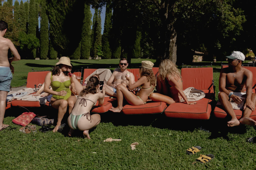 A Three-Day Destination Wedding in Tuscany 68 A group of people in swimsuits relax on orange lounge chairs outdoors, surrounded by green grass and tall trees, enjoying a sunny day.