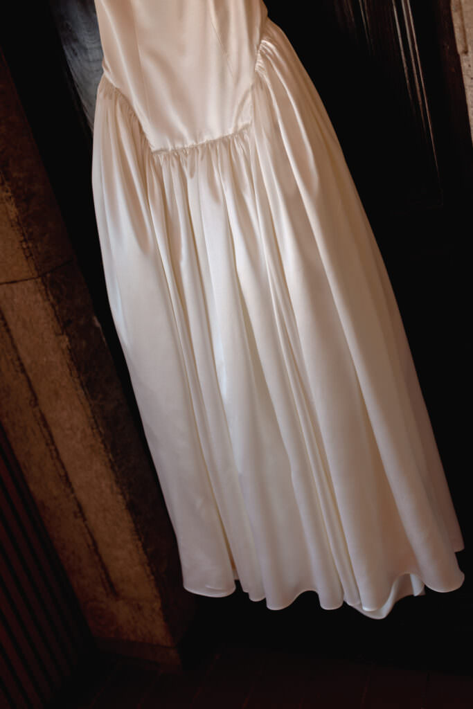 A Three-Day Destination Wedding in Tuscany 62 A white satin dress with a fitted bodice and gathered skirt hangs against a dark wooden door, with stone walls partially visible beside it.
