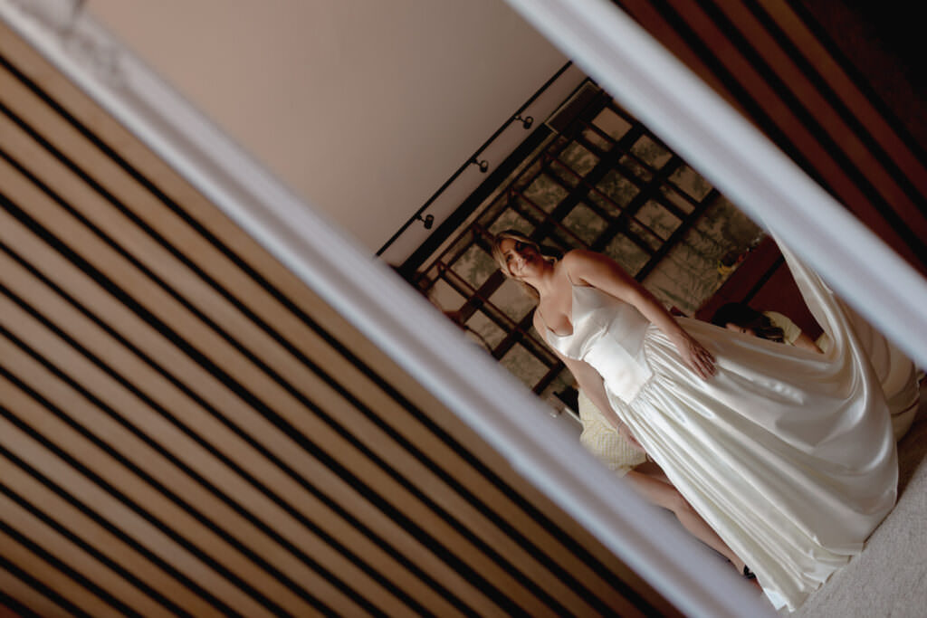 A Three-Day Destination Wedding in Tuscany 13 A bride in a white gown stands indoors, seen through a doorway at an angle. She is smiling slightly, and the room has a modern decor with wooden slats and framed pictures on the wall behind her.