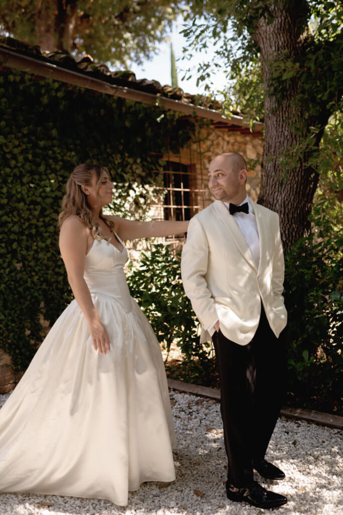 A Three-Day Destination Wedding in Tuscany 43 A bride in a white gown and a groom in a white tuxedo jacket stand outdoors on gravel, smiling at each other in front of a green ivy-covered wall and trees in daylight.