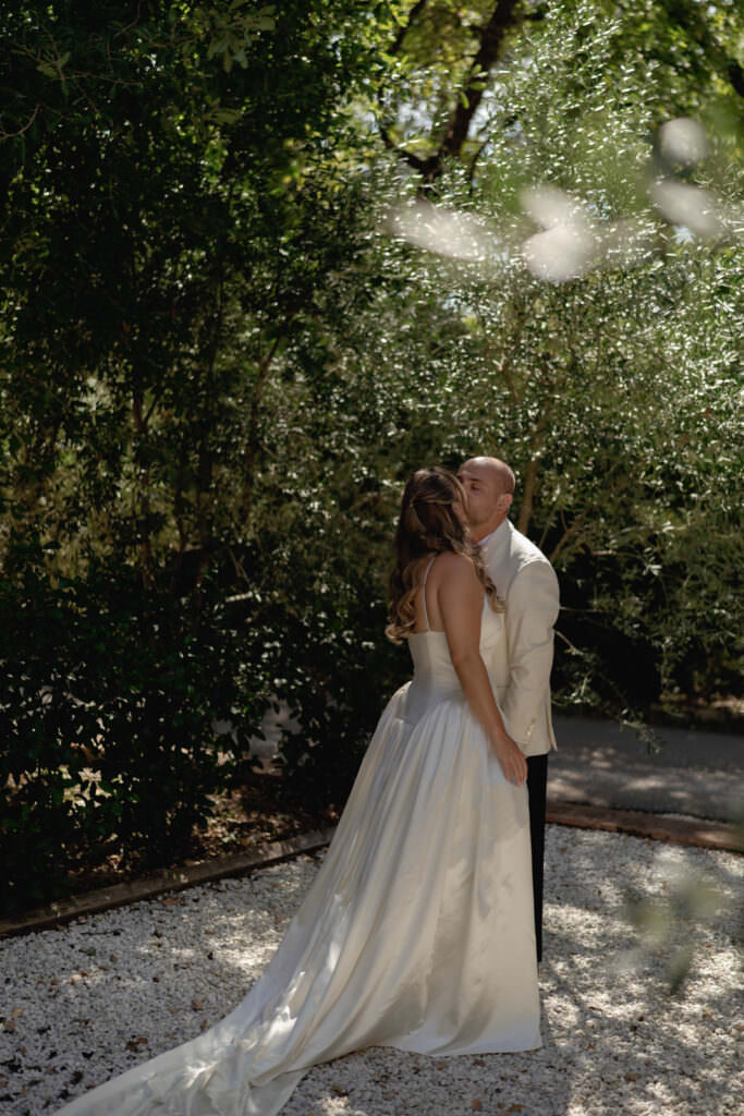 A Three-Day Destination Wedding in Tuscany 51 A bride in a white gown and a groom in a light-colored suit stand close together, sharing an intimate moment outdoors in a sunlit, leafy garden.