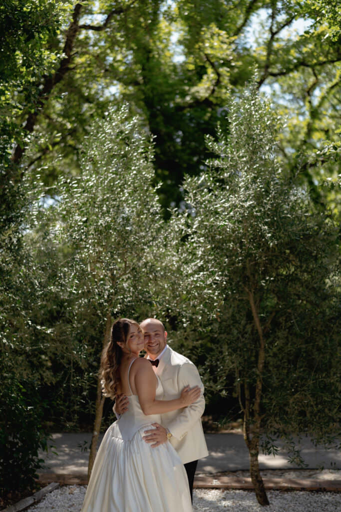 A Three-Day Destination Wedding in Tuscany 54 A smiling couple in wedding attire hugs outdoors, surrounded by lush green trees and sunlight filtering through the leaves. The bride wears a strapless white dress, and the groom wears a white jacket with a black bow tie.