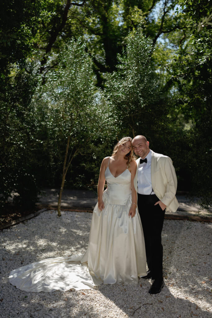 A Three-Day Destination Wedding in Tuscany 26 A bride in a white gown and a groom in a white jacket and black pants stand close together, smiling, surrounded by greenery and sunlight in an outdoor setting.