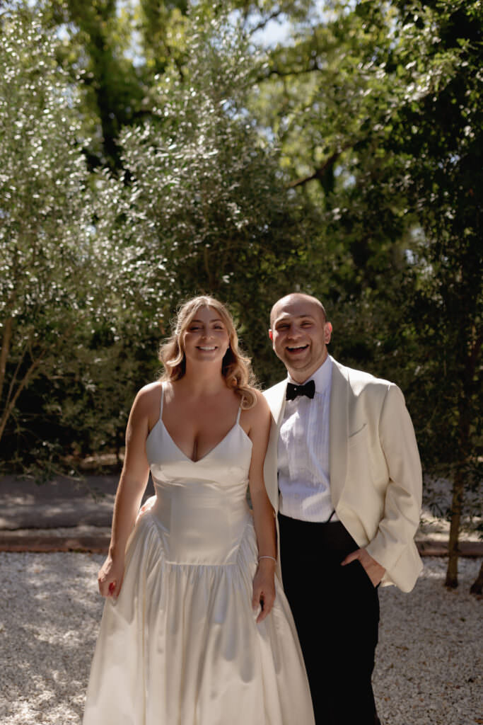 A Three-Day Destination Wedding in Tuscany 36 A smiling couple dressed in formal attire stands outdoors. The woman wears a satin white gown and the man wears a cream tuxedo jacket with a black bow tie. Sunlight filters through the trees in the background.