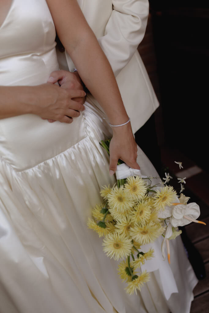 A Three-Day Destination Wedding in Tuscany 24 A bride in a satin white dress holds a bouquet of yellow flowers while her partner, dressed in white, gently embraces her from behind. Their faces are not visible, focusing on their hands and the bouquet.