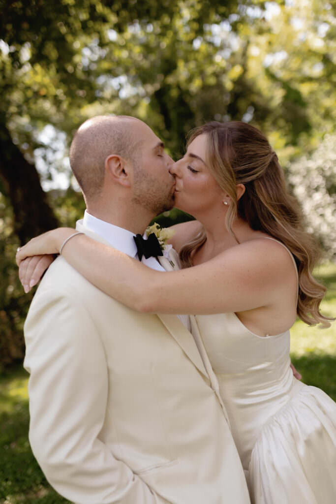 A Three-Day Destination Wedding in Tuscany 33 A bride and groom in cream-colored attire share a kiss outdoors under trees, embracing each other on their wedding day. Sunlight filters through the leaves, creating a romantic and intimate scene.