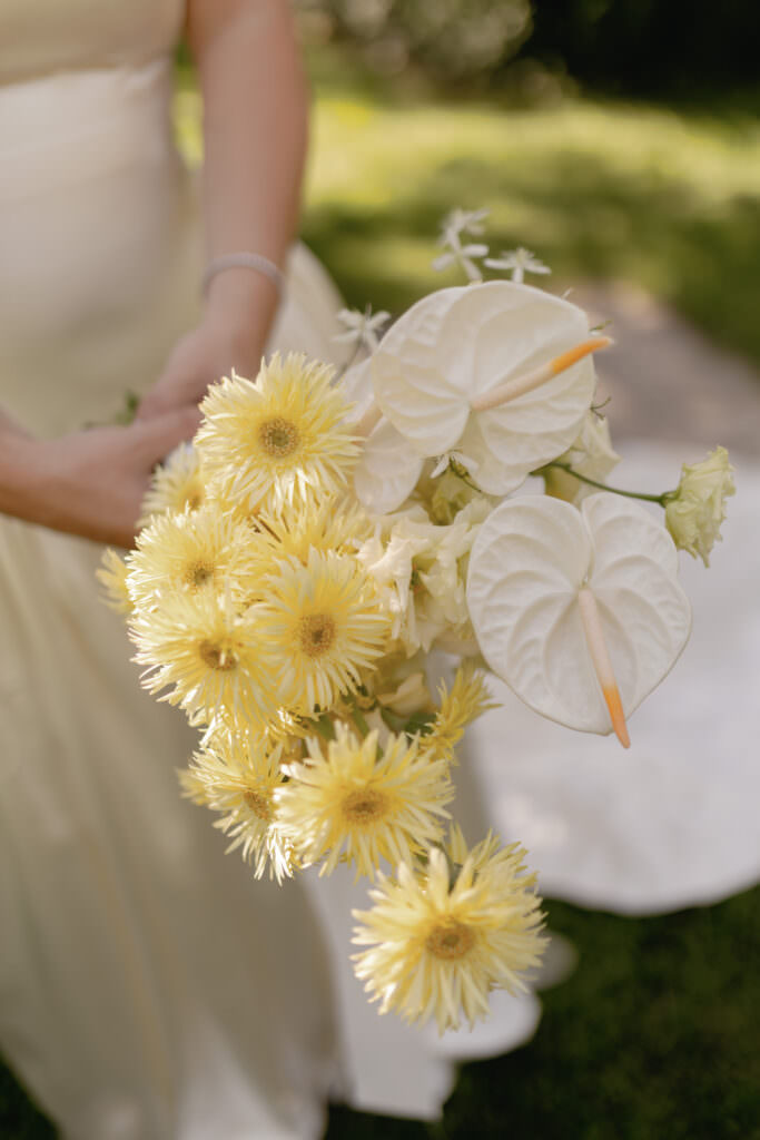 A Three-Day Destination Wedding in Tuscany 79 A person in a light-colored dress holds a bouquet of pale yellow chrysanthemums and white anthurium flowers outdoors, with greenery and sunlight in the background.