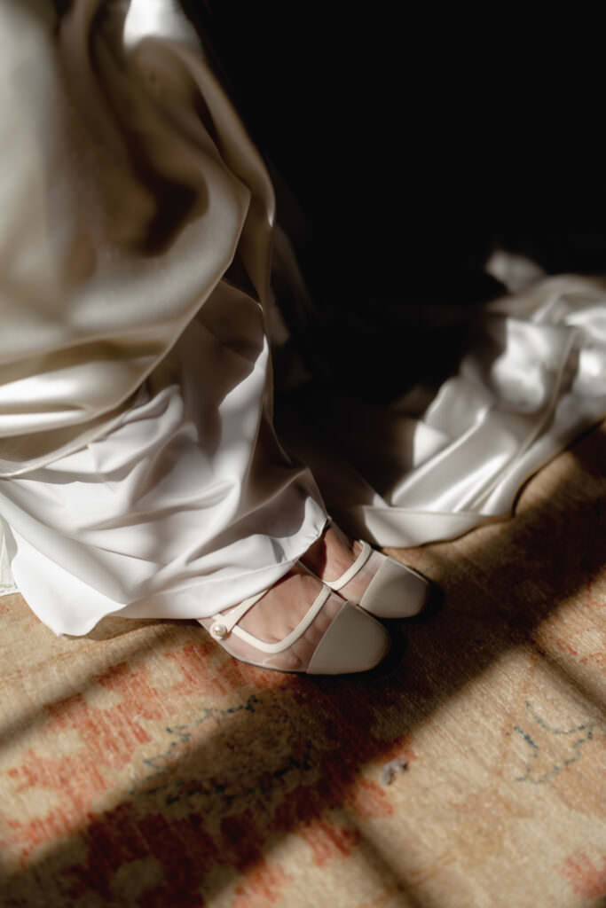 A Three-Day Destination Wedding in Tuscany 69 A close-up of a person wearing a white satin dress and white Mary Jane shoes, standing on a rug with sunlight casting shadows across the scene.