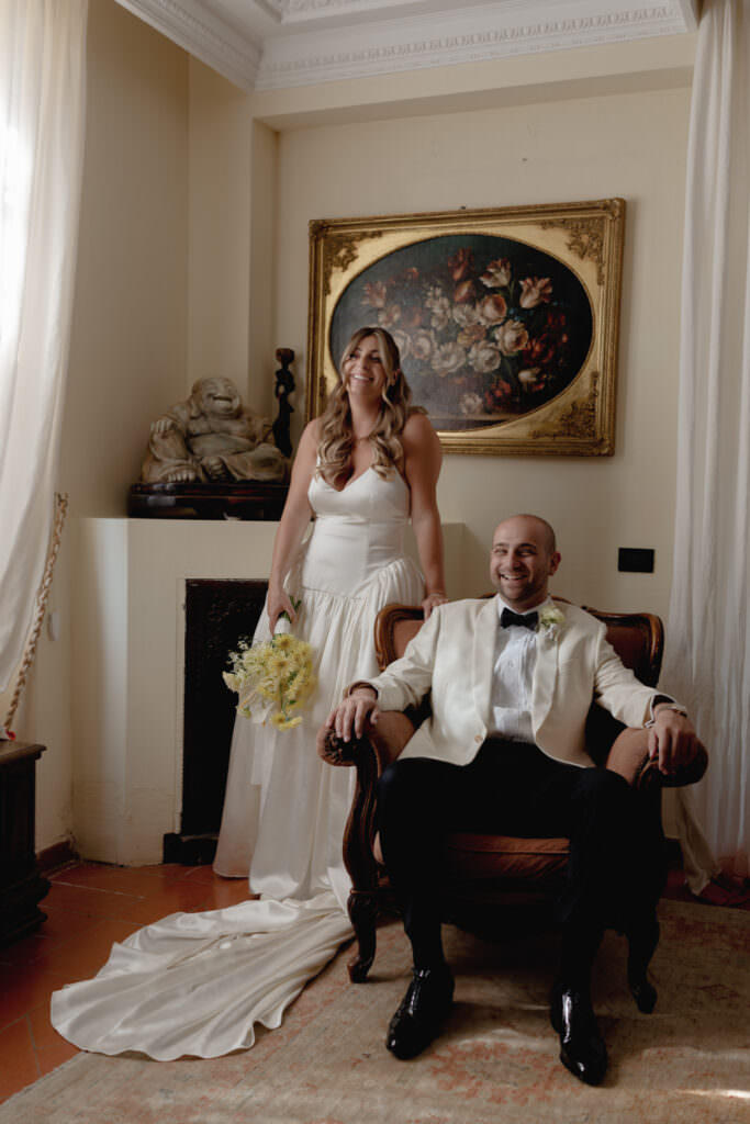 A Three-Day Destination Wedding in Tuscany 10 A bride in a white dress stands smiling behind a groom seated in an armchair, both indoors. The groom wears a white jacket and black pants. The room has elegant decor, with art and a statue in the background.