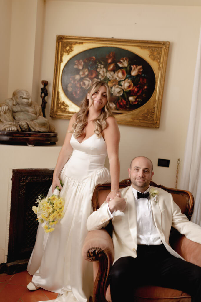 A Three-Day Destination Wedding in Tuscany 83 A bride in a white gown holding yellow flowers stands beside a seated groom in a white tuxedo jacket. They smile, holding hands, in a room with classic decor and a floral painting on the wall behind them.