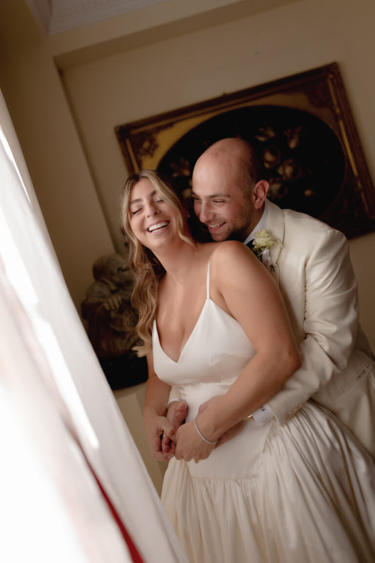 A Three-Day Destination Wedding in Tuscany 3 A smiling bride in a white dress stands in front of a groom in a cream suit as they embrace joyfully indoors, with soft light coming from the left and a framed picture in the background.