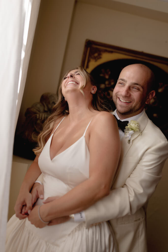 A Three-Day Destination Wedding in Tuscany 55 A newlywed couple smiles joyfully, standing close together. The bride wears a white dress and laughs, while the groom, in a white tuxedo jacket with a boutonnière, embraces her from behind near a softly lit window.
