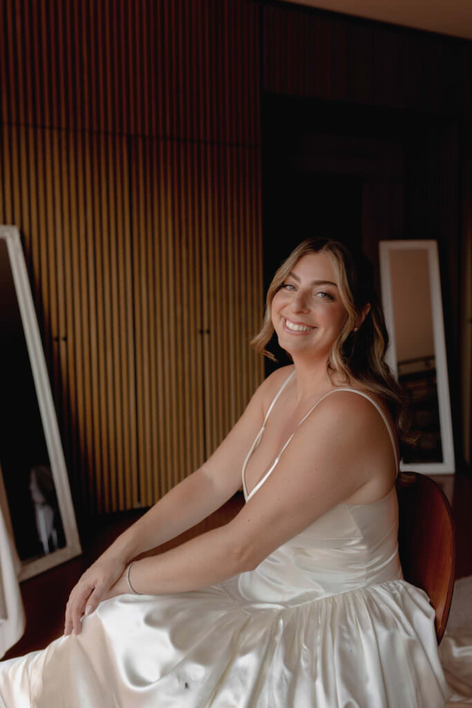 A Three-Day Destination Wedding in Tuscany 53 A woman in a white satin dress sits smiling, looking at the camera. She has wavy, shoulder-length hair and is indoors, with wooden paneling and mirrors in the background.