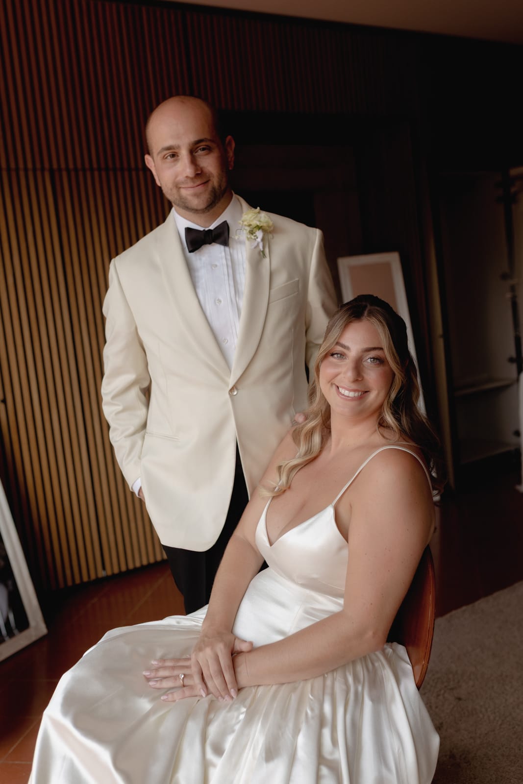 A smiling bride in a satin white dress sits while a groom stands beside her, wearing a cream-colored jacket and black bow tie. They pose indoors against a wooden wall, looking happy and relaxed.