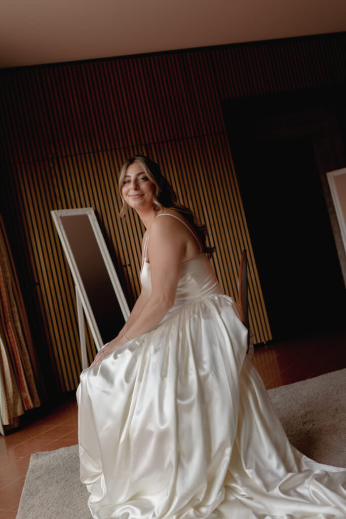 A Three-Day Destination Wedding in Tuscany 30 A bride in a satin wedding dress sits on a chair, smiling at the camera. She is indoors, with vertical wooden paneling and two standing mirrors in the background.