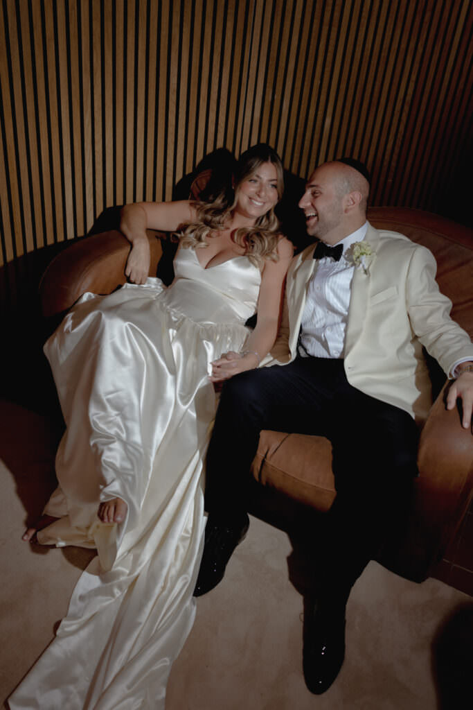A Three-Day Destination Wedding in Tuscany 90 A bride in a white satin gown and a groom in a cream jacket and black pants sit smiling together on a brown couch, holding hands and looking at each other happily.