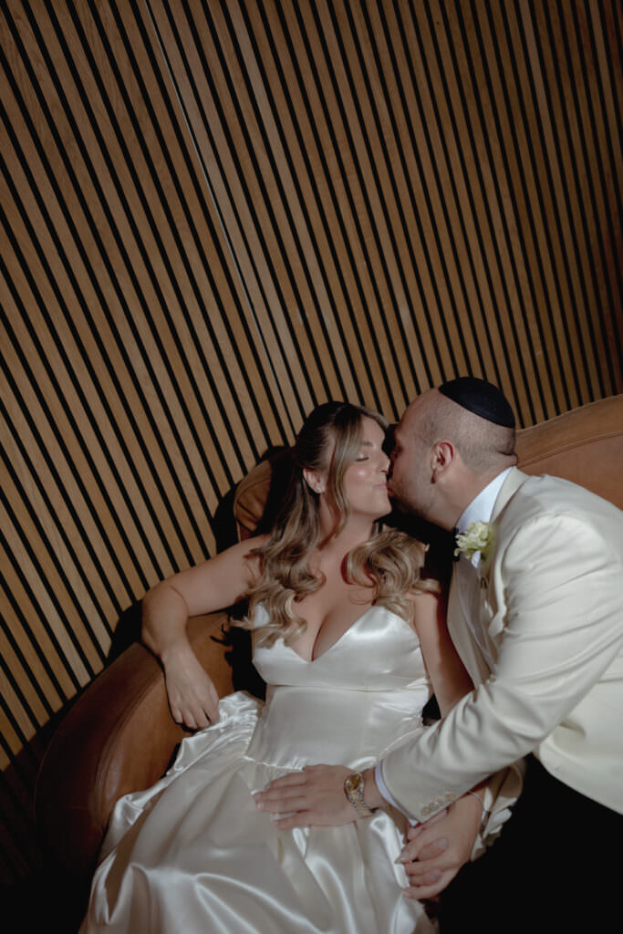 A Three-Day Destination Wedding in Tuscany 67 A bride in a white satin dress and a groom in a white tuxedo jacket and kippah share an intimate moment on a tan couch, leaning in for a kiss against a backdrop of wooden diagonal slats.